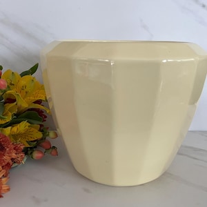 May include: A cream-colored, faceted ceramic vase. The vase has a wide opening and a geometric design. The vase is sitting on a white surface. A bouquet of yellow and orange flowers is next to the vase.