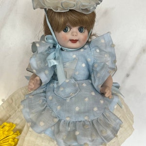 May include: A porcelain doll wearing a blue and white polka dot dress with a white lace trim and a white hat with a feather. The doll has brown hair, blue eyes, and rosy cheeks.