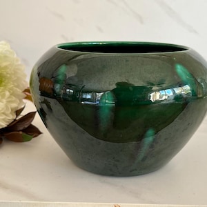 May include: A round, deep green ceramic bowl with a smooth, glossy finish. The bowl features a wide, open top and a curved, bulbous body. The dark green glaze has subtle variations in tone, reflecting light across its polished surface.