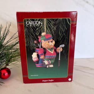 May include: A Carlton Cards Christmas ornament featuring a bear dressed as a golfer. The bear wears a red and green sweater and hat, holding a golf club. The ornament is in a red box with the text "Dapper Duffer" and "1998".