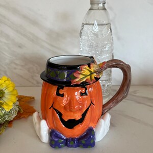 May include: A ceramic Halloween-themed mug shaped like a smiling pumpkin with a black hat and purple bow tie. The mug has a brown handle and white hands. A clear water bottle is behind the mug.