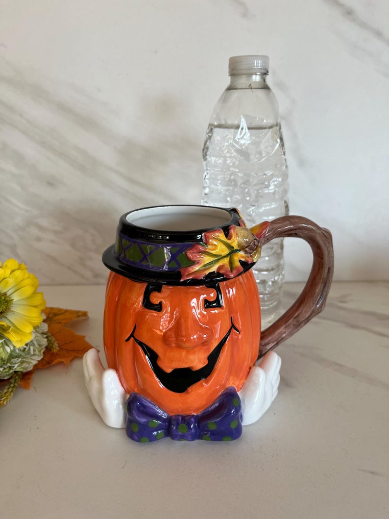May include: A ceramic Halloween-themed mug shaped like a smiling pumpkin with a black hat and purple bow tie. The mug has a brown handle and white hands. A clear water bottle is behind the mug.