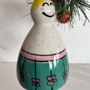 May include: A ceramic Christmas ornament shaped like a smiling figure with a yellow head and a white body decorated with pink polka dots. The figure has a green skirt with black vertical stripes and pink flowers. A gold string is attached for hanging.