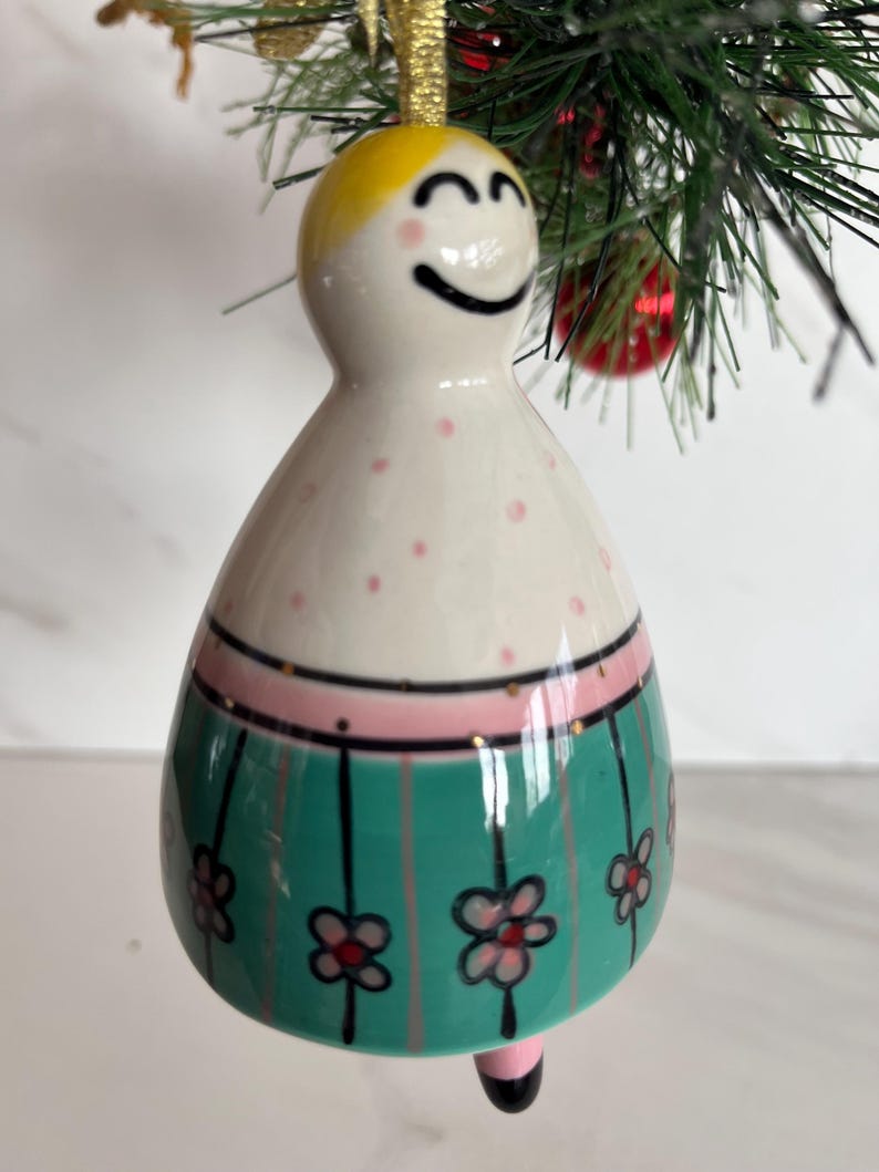 May include: A ceramic Christmas ornament shaped like a smiling figure with a yellow head and a white body decorated with pink polka dots. The figure has a green skirt with black vertical stripes and pink flowers. A gold string is attached for hanging.