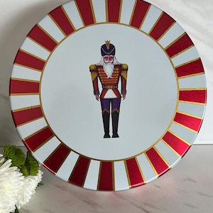 May include: Circular tin with a white base and a red and gold striped border. The centre features a detailed illustration of a nutcracker soldier in a red and gold uniform. The tin is likely for storage.