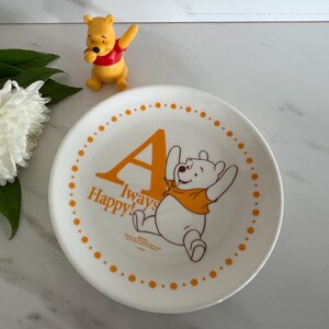 May include: A white ceramic plate with a Winnie the Pooh design. The plate features the letter "A", the words "Always Happy!", and an illustration of Winnie the Pooh. A small Winnie the Pooh figurine is also present.