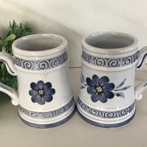 May include: Two white ceramic mugs with blue floral designs and blue and white swirl patterns around the top and bottom.