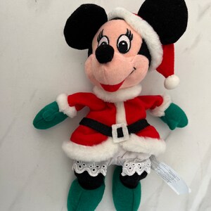 Puede incluir: Un juguete de peluche de Minnie Mouse vestida con un traje de Pap&aacute; Noel rojo y blanco con un gorro verde. Lleva un ribete de encaje blanco en la falda y un cintur&oacute;n negro.
