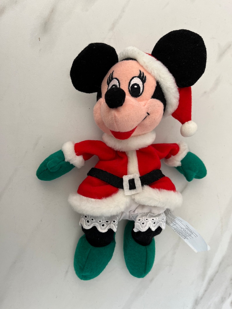 Puede incluir: Un juguete de peluche de Minnie Mouse vestida con un traje de Pap&aacute; Noel rojo y blanco con un gorro verde. Lleva un ribete de encaje blanco en la falda y un cintur&oacute;n negro.