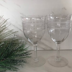 May include: Two clear wine glasses with etched horizontal bands. The glasses have a classic design with a rounded bowl and a slender stem. The glasses are displayed on a white surface, with a sprig of green pine needles and red ornaments in the background.