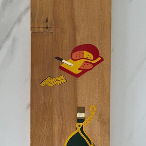 May include: Wooden serving board featuring painted cheese, crackers, and a wine bottle. The board has a handle and is made of light-coloured wood. The cheese is red and yellow, the crackers are yellow, and the wine bottle is yellow and green.