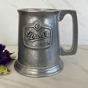 May include: A pewter beer mug with a handle. The mug features the Stroh's Beer logo in a rectangular emblem. The mug is cylindrical with a flared base and a sturdy handle. Purple flowers and green leaves are in the foreground.