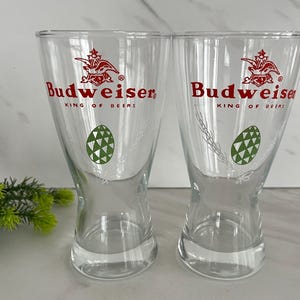 May include: Two clear Budweiser beer glasses. Each glass has the Budweiser logo in red, with the words "KING OF BEERS" below. A green and white geometric design is also featured on each glass.
