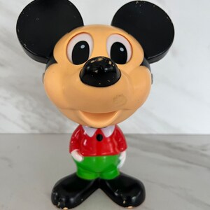May include: A vintage plastic Mickey Mouse figurine with black ears, large eyes, a red shirt, and green pants.