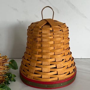 May include: A bell-shaped woven basket with a light brown finish, featuring a small metal loop handle at the top. The base is accented with a red and green painted rim, secured with small metal tacks around the circumference.