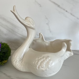 May include: A cream-coloured ceramic swan planter with an outstretched neck and open basin. The swan has detailed feather patterns and a glossy finish. The planter is designed to hold plants or flowers, adding a decorative touch to any space.