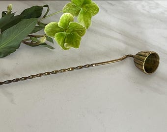 May include: A long brass cocktail spoon featuring a twisted handle and a small, fluted, cup-shaped bowl at the end. The metal has a warm, aged patina, and the design includes a distinct bend connecting the handle to the rounded, ribbed bowl.