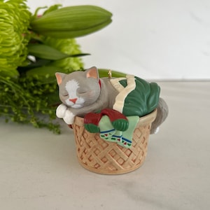 May include: A gray and white cat ornament sleeping in a wicker basket filled with green and red Christmas stockings.