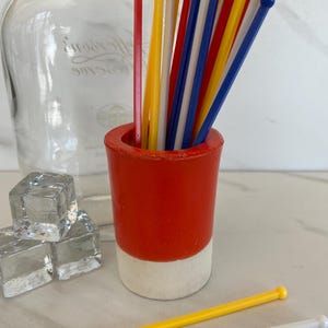 May include: A red and white cup holding a collection of cocktail stirrers in various colours. The stirrers are red, yellow, white, and blue. A clear glass bottle and ice cubes are in the background. The cup is made of ceramic or a similar material.