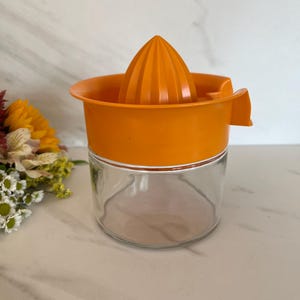 May include: An orange citrus juicer with a clear glass base. The juicer has a conical reamer and a wide rim with a spout. The design is simple and functional, perfect for extracting fresh juice. The juicer is likely made of plastic and glass.