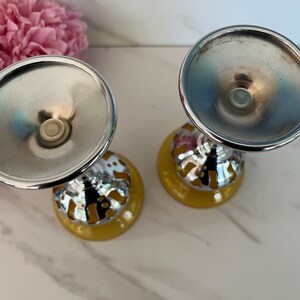 May include: Two decorative goblets with a silver-colored bowl and stem, and a yellow base. The stem has a decorative pattern with gold and pink accents. The goblets are set against a background of pink flowers.