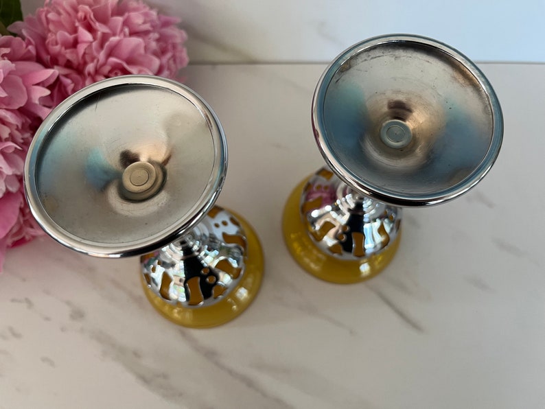 May include: Two decorative goblets with a silver-colored bowl and stem, and a yellow base. The stem has a decorative pattern with gold and pink accents. The goblets are set against a background of pink flowers.