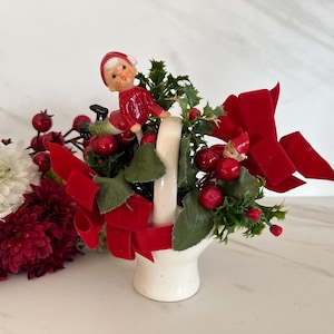 May include: A white ceramic basket filled with red berries, green holly leaves, and red velvet bows. Two small ceramic elves are perched on the rim of the basket.