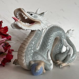 May include: A porcelain dragon figurine featuring a glossy light blue and grey finish with a white underbelly. The dragon has an open mouth with a pink tongue, sharp teeth, and is holding a small blue sphere with its front clawed feet.