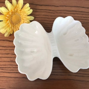 May include: A white, bow-shaped serving dish with scalloped edges. The dish is made of a glossy material and has two separate compartments. A yellow flower with a green stem is next to the dish.