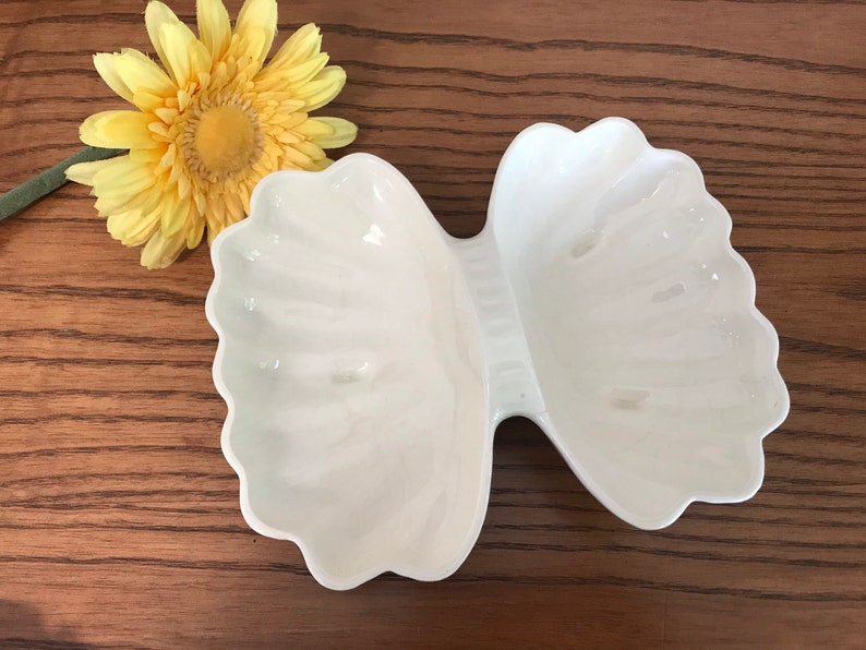 May include: A white, bow-shaped serving dish with scalloped edges. The dish is made of a glossy material and has two separate compartments. A yellow flower with a green stem is next to the dish.