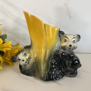 May include: A ceramic vase shaped like a tree stump with a yellow and green gradient finish. Two small bear figures are attached to the base, one on each side of the central opening. The bears are painted in shades of gray and black with detailed faces.
