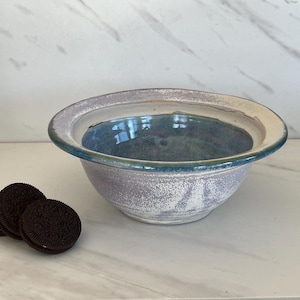 May include: A handmade ceramic bowl with a wide rim and a speckled, textured surface. The bowl features a light purple exterior, a blue interior, and a cream-colored rim. Three chocolate sandwich cookies are next to the bowl.