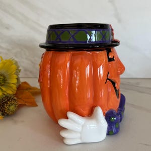May include: Orange ceramic pumpkin-shaped container with a black hat, a painted face, and a white glove. The hat has a green and purple design. The container is likely for Halloween or autumn decor.