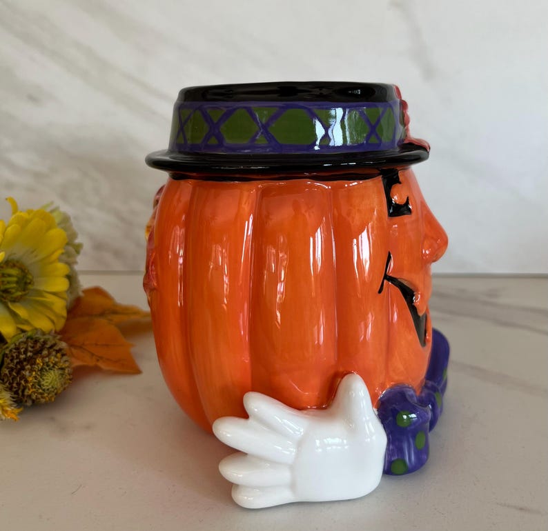 May include: Orange ceramic pumpkin-shaped container with a black hat, a painted face, and a white glove. The hat has a green and purple design. The container is likely for Halloween or autumn decor.