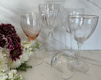 May include: Four clear glass wine glasses, one with a gold rim, on a white marble surface.