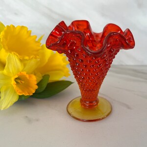 May include: A red glass vase with a ruffled top and a textured, dotted body. The vase has a yellow base.