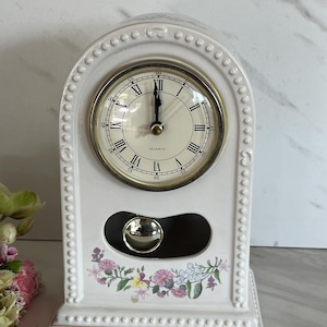 May include: An arched white ceramic mantel clock with a beaded border and a floral decal at the base. The clock face features black Roman numerals, a gold-toned bezel, and the word QUARTZ. A silver-toned pendulum is visible through a cutout.