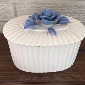 May include: A white ceramic trinket box with a lid, featuring a ribbed design and a blue rose accent on top. The box is round with vertical ridges and sits on a wooden surface. A decorative piece for storage.