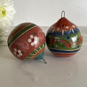 May include: Two hand-painted wooden spinning tops. One is red with green stripes and white floral accents. The other is red, green, and blue with white and red painted designs. Both have metal tips and are on a white surface.