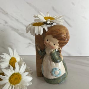 May include: A ceramic figurine of a person with brown hair leaning against a brown brick-patterned pillar that serves as a vase. The person wears a white apron with blue flower accents over a green garment. Two white daisies with yellow centers are placed in the pillar vase, with additional daisies resting nearby.