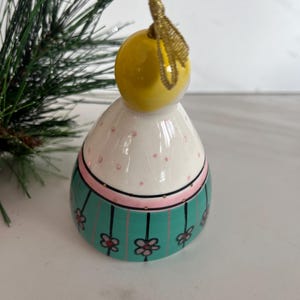 May include: A ceramic Christmas ornament shaped like a stylized figure. The ornament has a yellow head, a white body with pink polka dots, and a teal band with floral accents. A gold ribbon is attached for hanging.