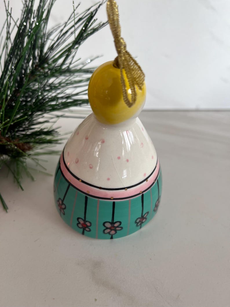 May include: A ceramic Christmas ornament shaped like a stylized figure. The ornament has a yellow head, a white body with pink polka dots, and a teal band with floral accents. A gold ribbon is attached for hanging.