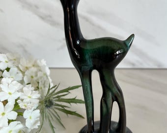 May include: A ceramic deer figurine with a glossy, dark green and black glaze. The deer has a slender neck, upright ears, and a rounded base. The figurine is displayed with white flowers and greenery.