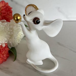 May include: A white ceramic mouse figurine with large brown eyes and a gold-colored ball. The mouse is holding the ball in its raised paw. The figurine has a gold-colored rim around its head. Red and white flowers are in the background.