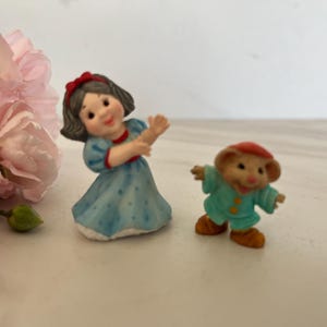 May include: Figurines of a girl in a blue dress with a red bow and a mouse in a green outfit. The girl figurine has her arms raised. The mouse figurine has its arms out. Pink flowers are in the background.