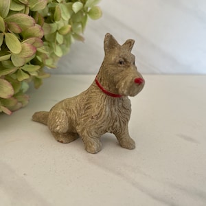 May include: A small, gray, ceramic figurine of a Scottish Terrier dog wearing a red collar. The dog is sitting with its head turned to the right.