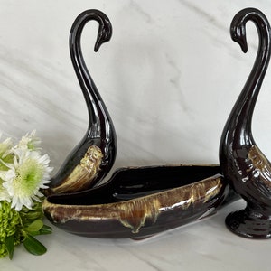 May include: A brown and black ceramic swan planter with a brown and yellow glaze. The planter is shaped like a swan with its neck curved up. There are two swans in the image, one on each side of a brown and yellow ceramic bowl.