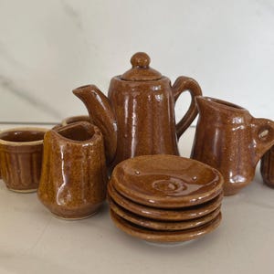 May include: A miniature brown ceramic tea set, including a teapot, creamer, sugar bowl, cups, and saucers. The set has a glossy finish and is arranged on a white surface. A yellow flower is in the background.