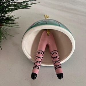 May include: A ceramic Christmas ornament shaped like a bell, with a teal and white exterior. Inside, pink legs with black ballet shoe detailing extend downwards. The ornament has a gold-coloured loop for hanging.