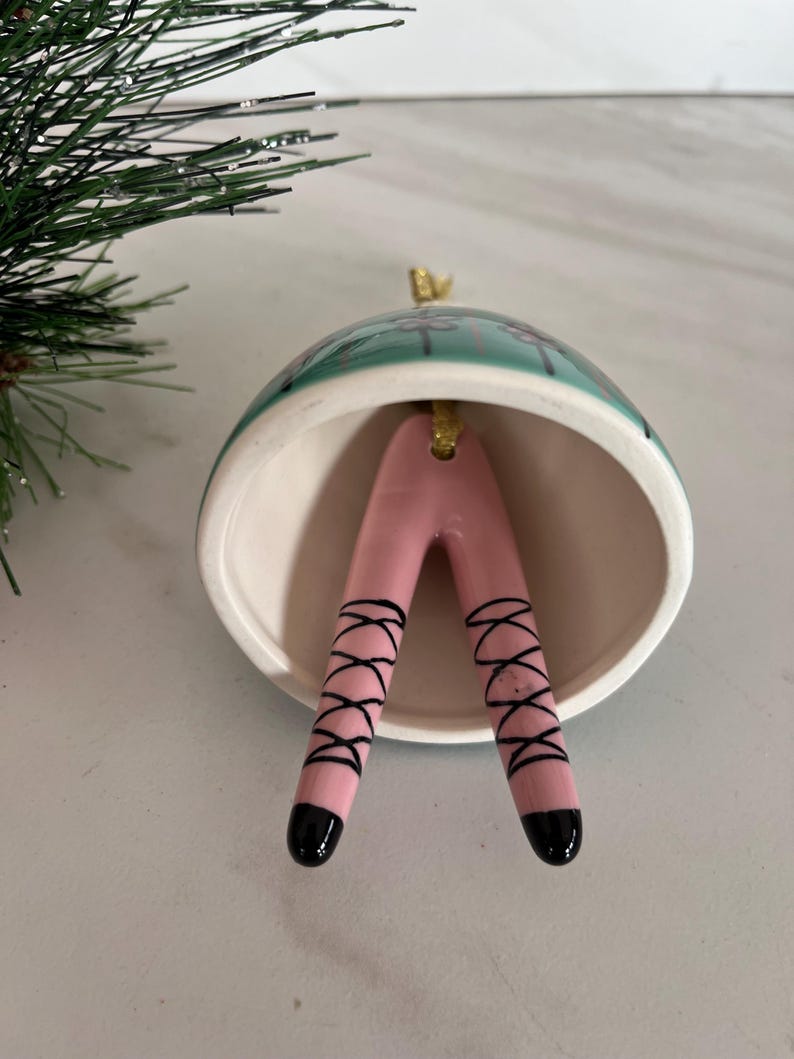 May include: A ceramic Christmas ornament shaped like a bell, with a teal and white exterior. Inside, pink legs with black ballet shoe detailing extend downwards. The ornament has a gold-coloured loop for hanging.